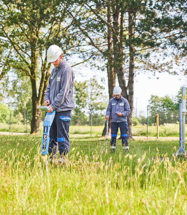 Zwei Männer, auf offenen Feld, tragen Schutzhelme und nehmen Messungen vor.